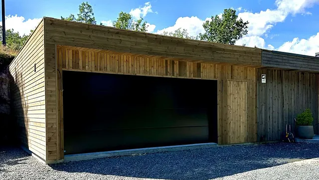 En garasje med trefasade og stor, svart garasjedør, fotografert i solskinn.