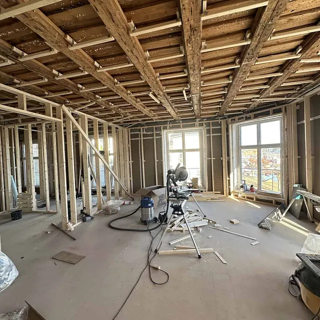 Innendørs byggeplass med synlige bjelker, verktøy på gulvet og vinduer.