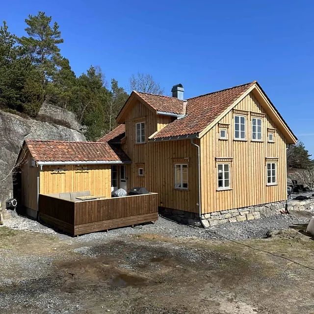 Ferdig kledd hus med et påbygg og platting mot en fjellvegg.
