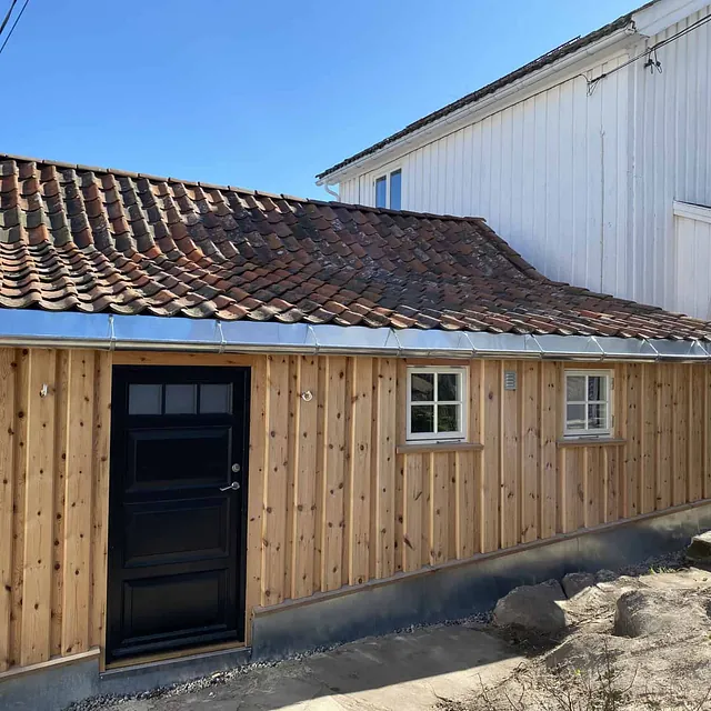 Et tilbygg i treverk, med ny takrenne og røde takstein, ved siden av et hvitt hus.