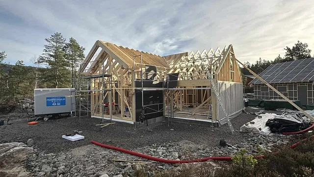 Et hus under bygging i skogen, med synlig reisverk og stillas.