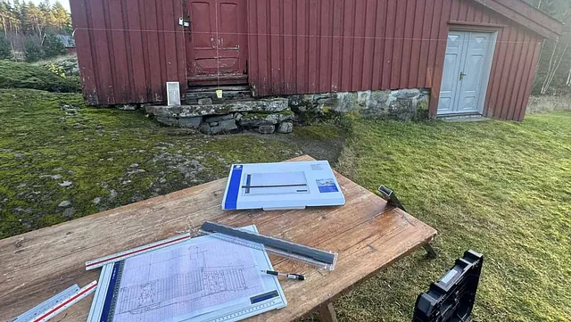 Et gammelt, rødt hus med synlige takbjelker og takstein, med plantegninger på et bord foran.