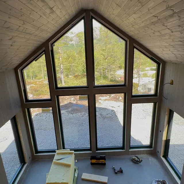 Stort rom med vinduer, skråtak og synlige bjelker, med byggematerialer på gulvet.