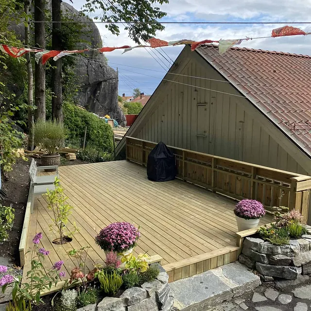 En treplatting med blomster i forgrunnen og et hus med takstein bak.