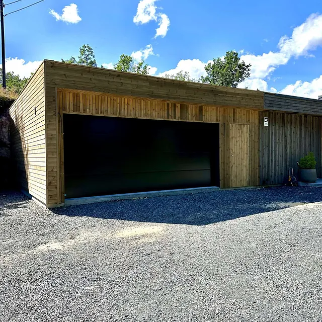 En garasje med trefasade og stor, svart garasjedør i solskinn.